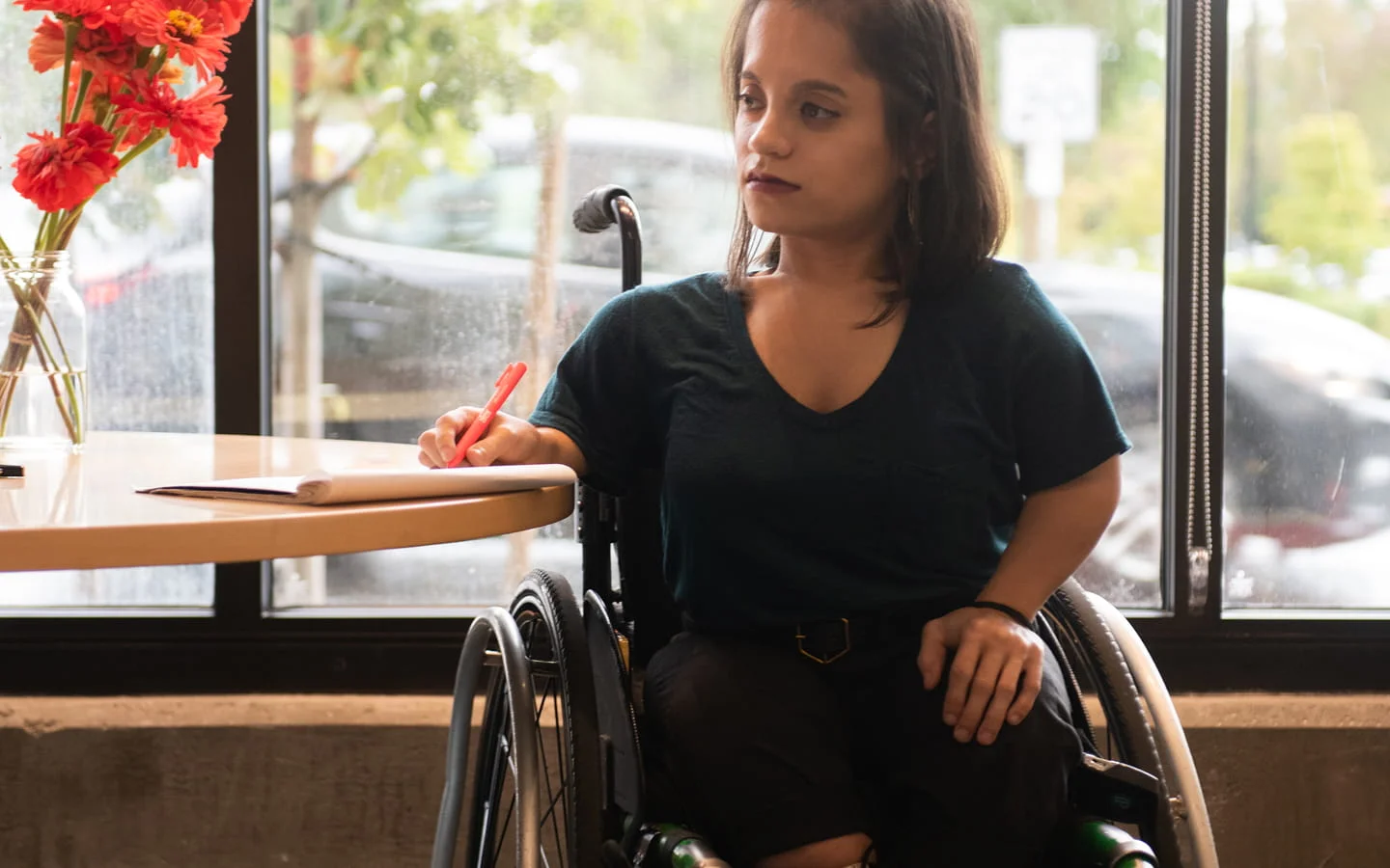 A disabled woman using a wheelchair sits at a round cafe table near a sunlit window, writing with a red marker and looking up